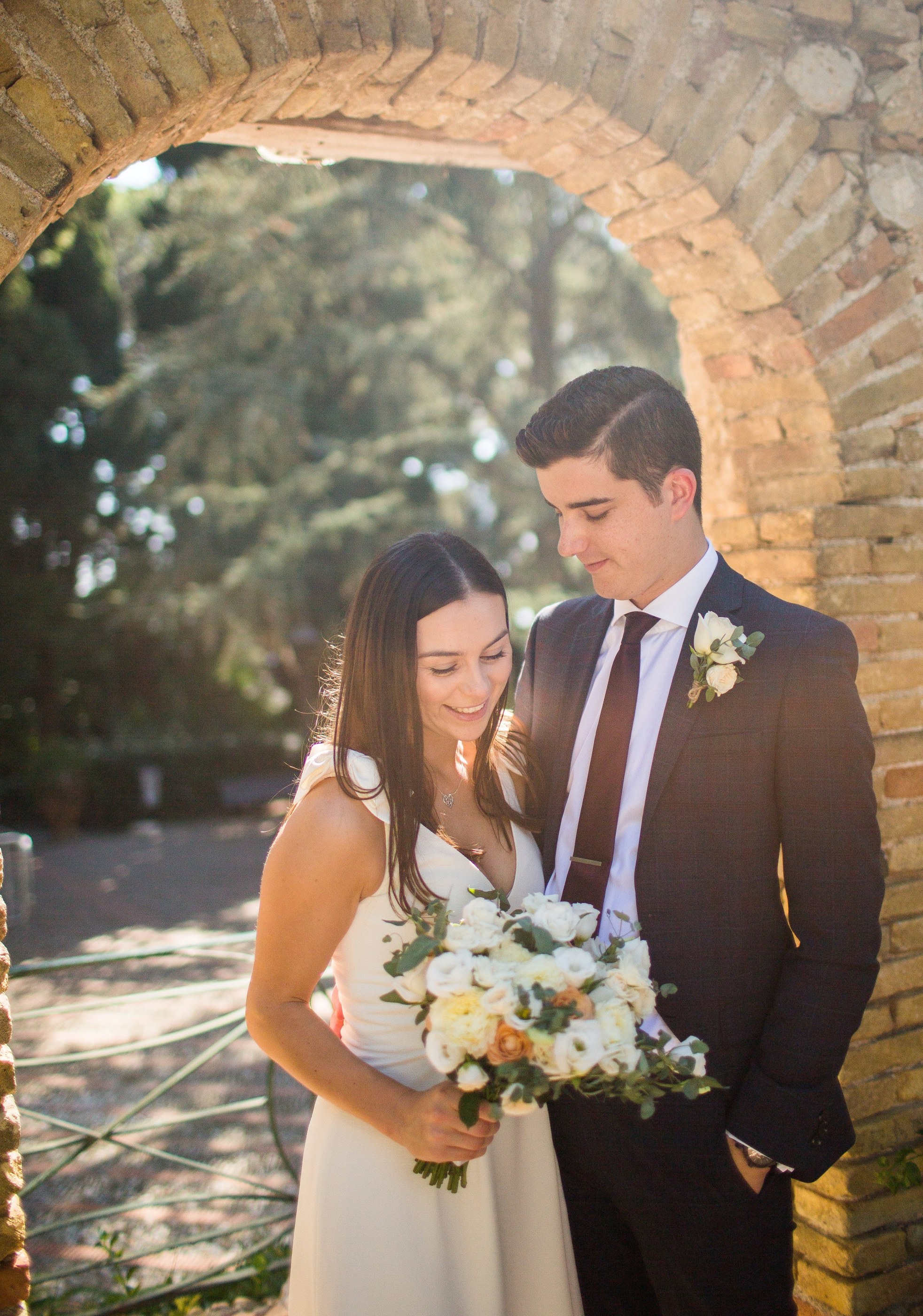 Michel and Heather standing close together beneath an old brick archway in Taormina, Heather in a white dress holding a large white and peach rose bouquet, Michel in a dark checked suit with a burgundy tie