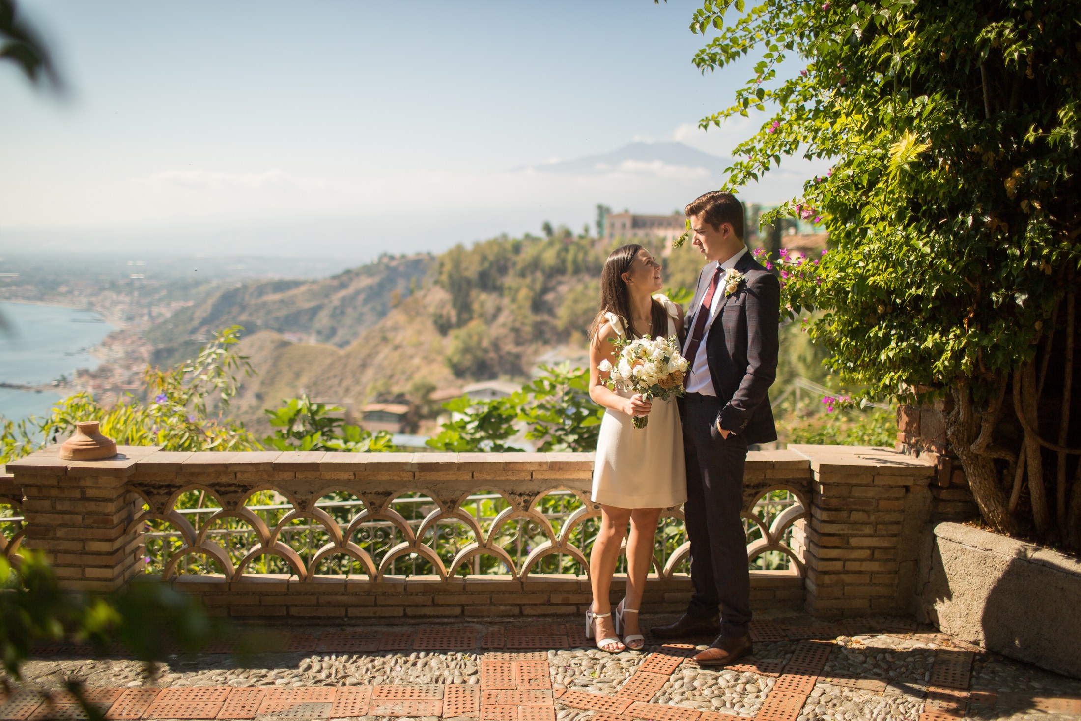 Michel and Heather walking hand in hand across the black and white checkerboard paving of Piazza IX Aprile in Taormina, Heather in a short white dress carrying a peach and white bouquet