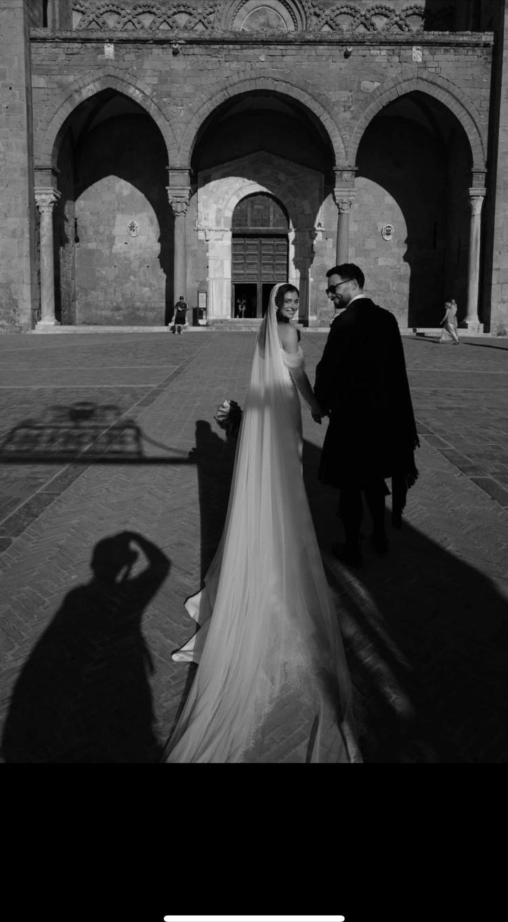Black and white photograph of a Sicilian wedding couple walking hand in hand across a piazza, the bride long veil trailing behind