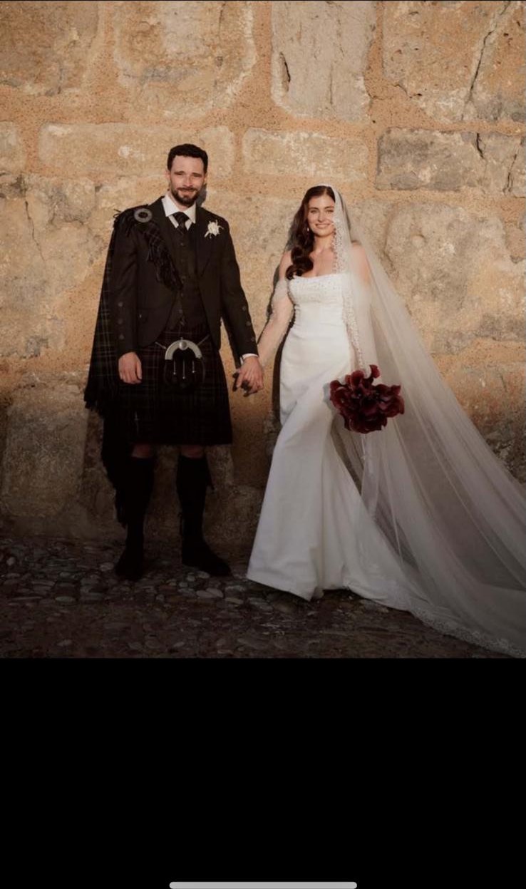 Wedding in Sicily — a bride in a white gown and long lace veil beside her groom in full Scottish highland dress against a golden Sicilian stone wall