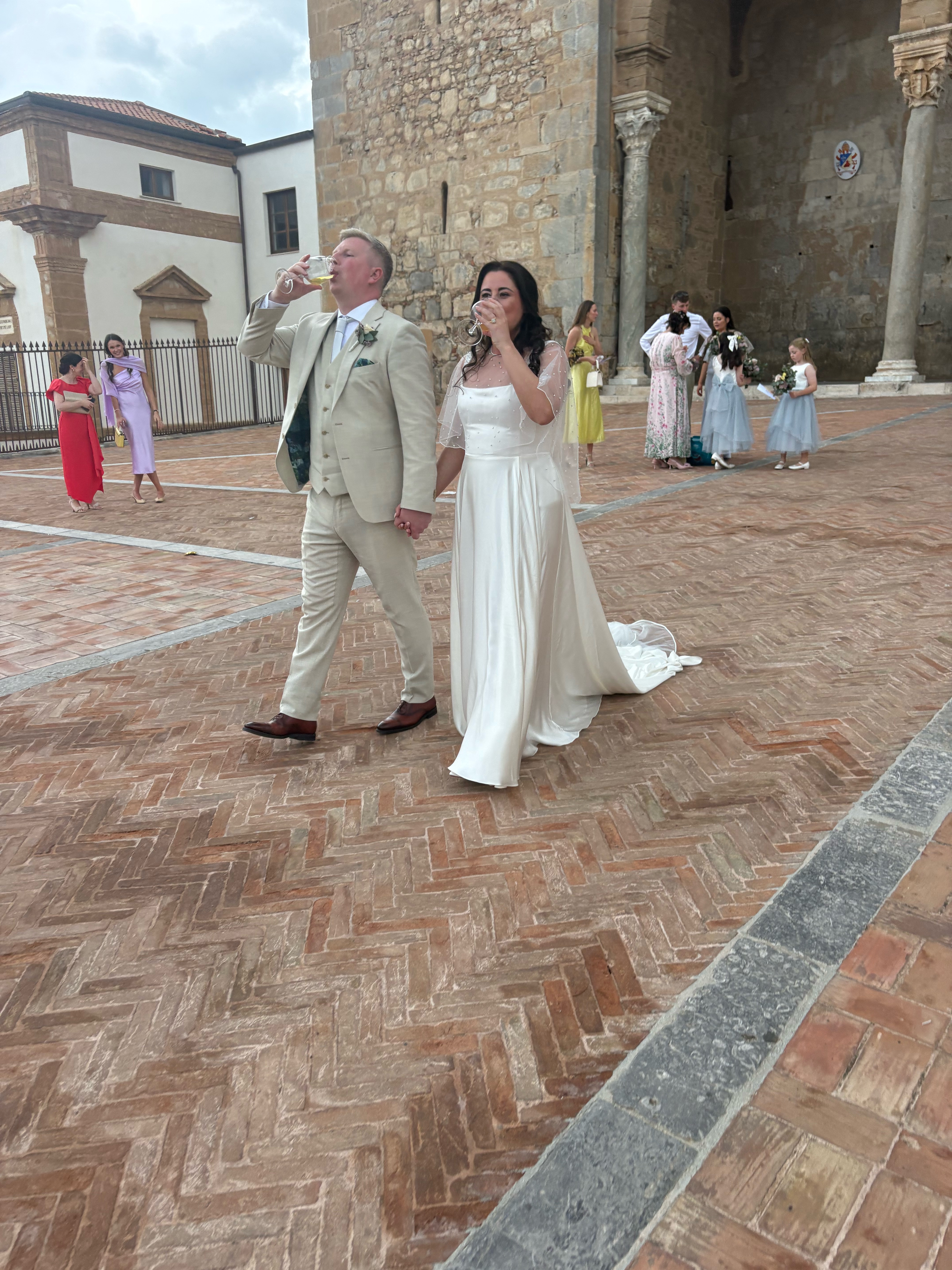 Gráinne and Phil walking hand in hand across the piazza outside Cefalù cathedral, each with a celebratory drink, as guests and flower girls follow behind