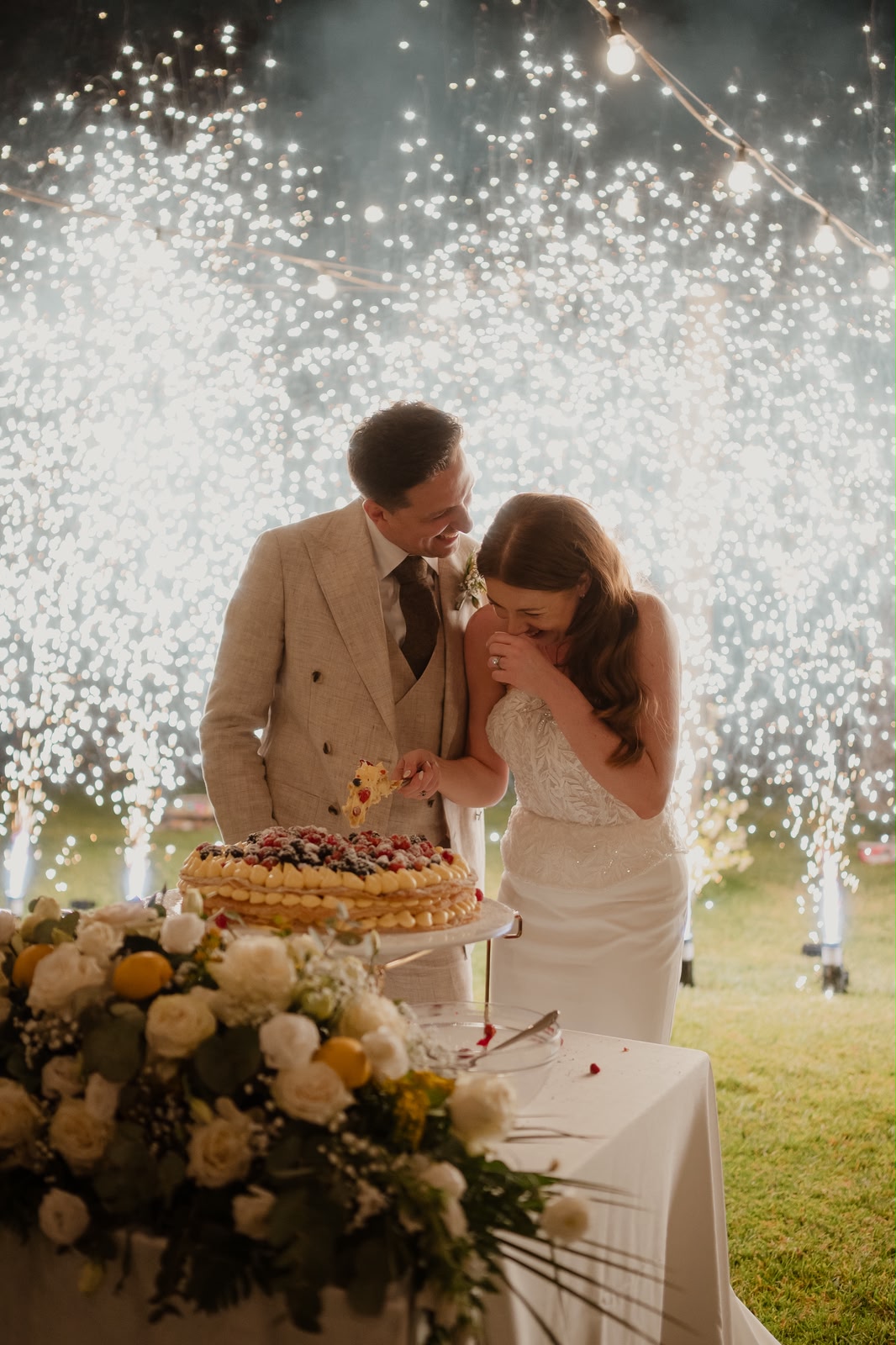 Jessica and Seb cutting their wedding cake as sparklers light up the evening