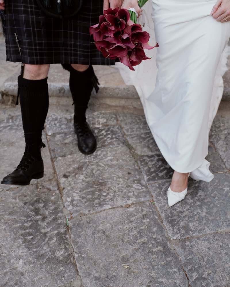 Sicilian wedding detail — a tartan kilt and brogues beside the bride shoes and a deep burgundy calla lily bouquet on cobbled stone