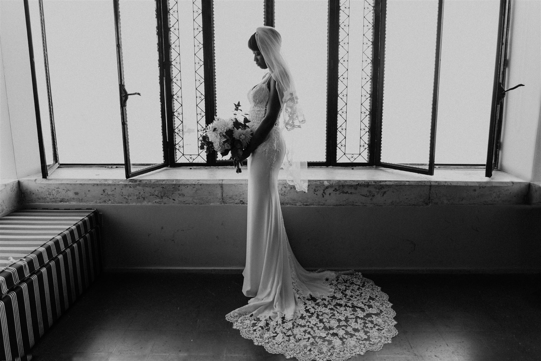 Ritratto editoriale in bianco e nero di Kysha in un abito di pizzo aderente e un lungo velo, con il bouquet tra le mani, accanto alle alte finestre a vetri piombati di Villa Mon Repos