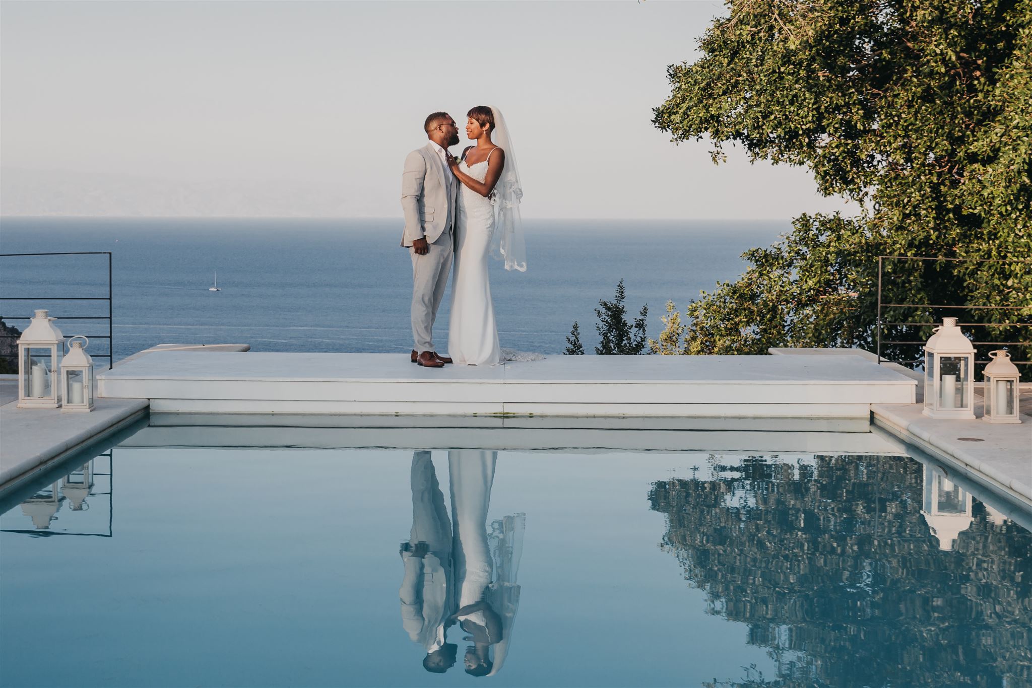Kysha e Shem insieme sul bordo di una piscina a specchio a Villa Mon Repos, con il Mar Ionio calmo che si estende fino all’orizzonte dietro di loro