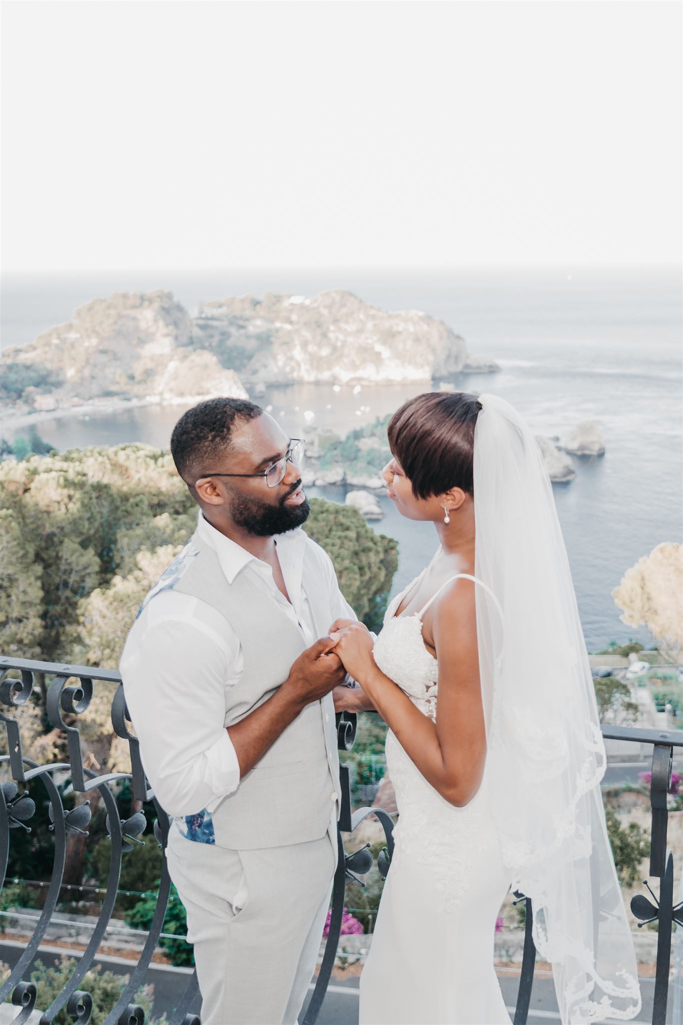 Kysha e Shem si tengono per mano su un balcone in ferro battuto di una villa a Taormina, con Isola Bella e la baia visibili sullo sfondo