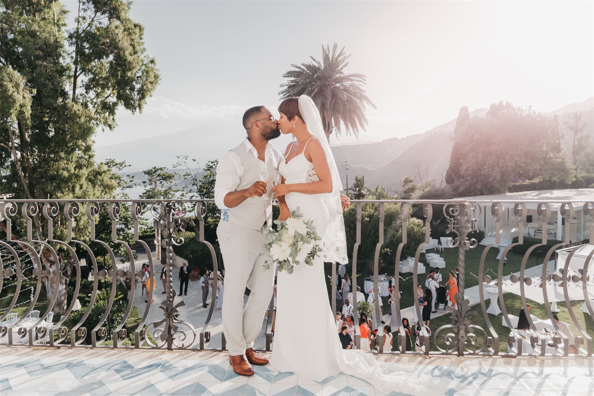 Bride and groom sharing a kiss with champagne in hand on an ornate wrought-iron balcony overlooking the Sicilian coast at golden hour