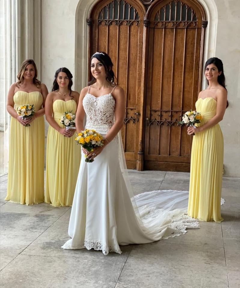 Bride with her three bridesmaids in pale-yellow gowns holding bouquets before a grand carved wooden church door in Sicily