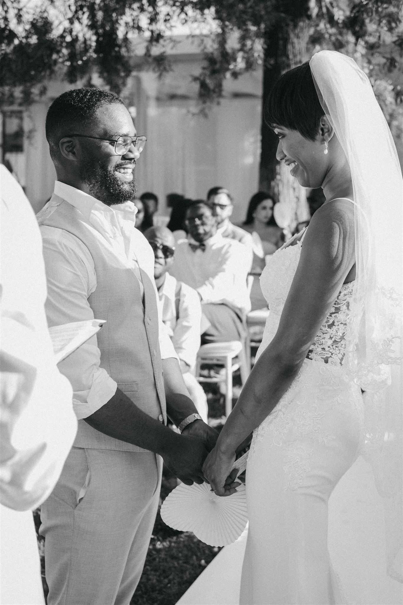 Black and white photograph of a bride and groom holding hands and smiling at each other during their outdoor Sicilian wedding ceremony as guests look on