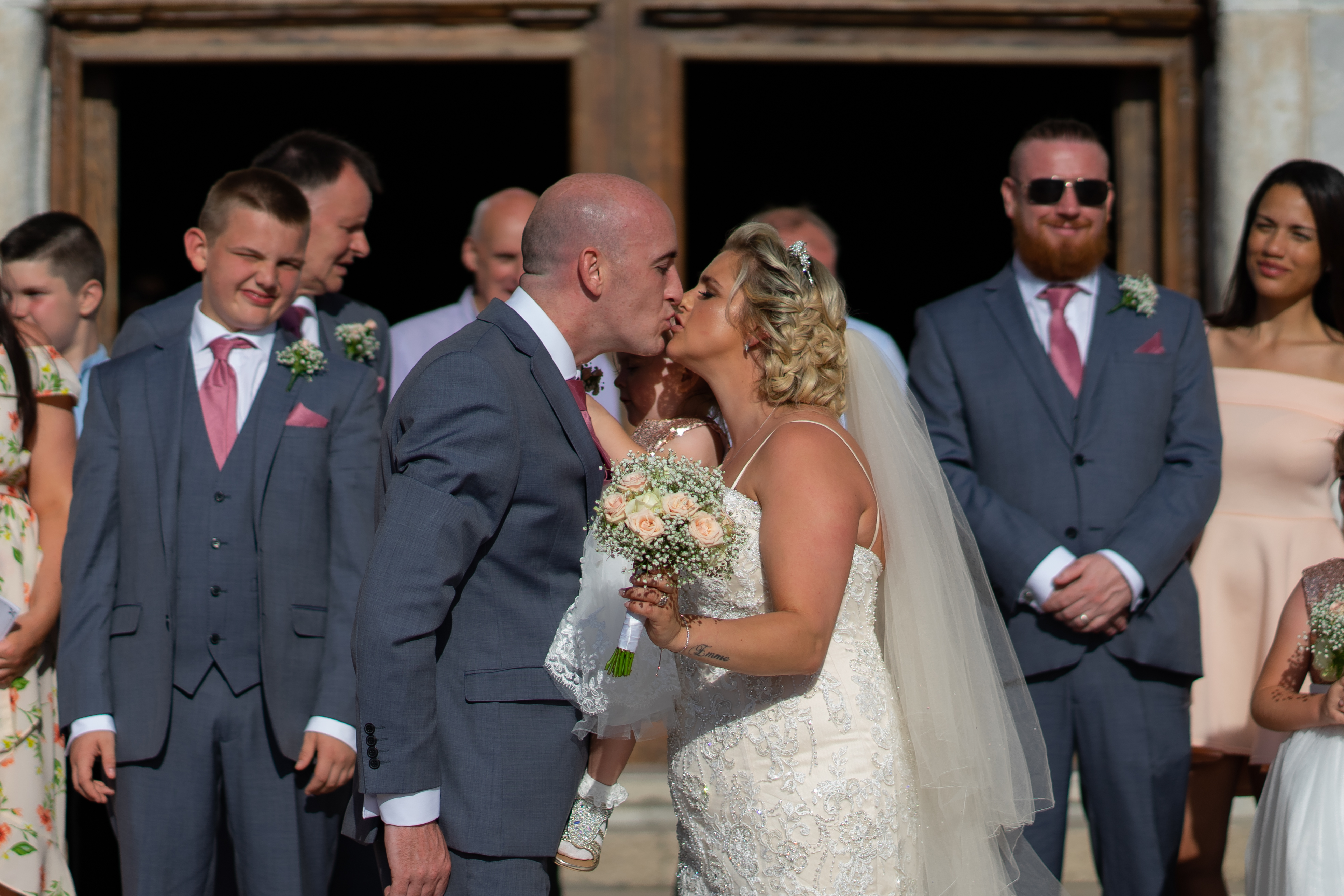 Bride and groom sharing their first kiss in a sunlit stone arched church doorway as their wedding party looks on in pink ties
