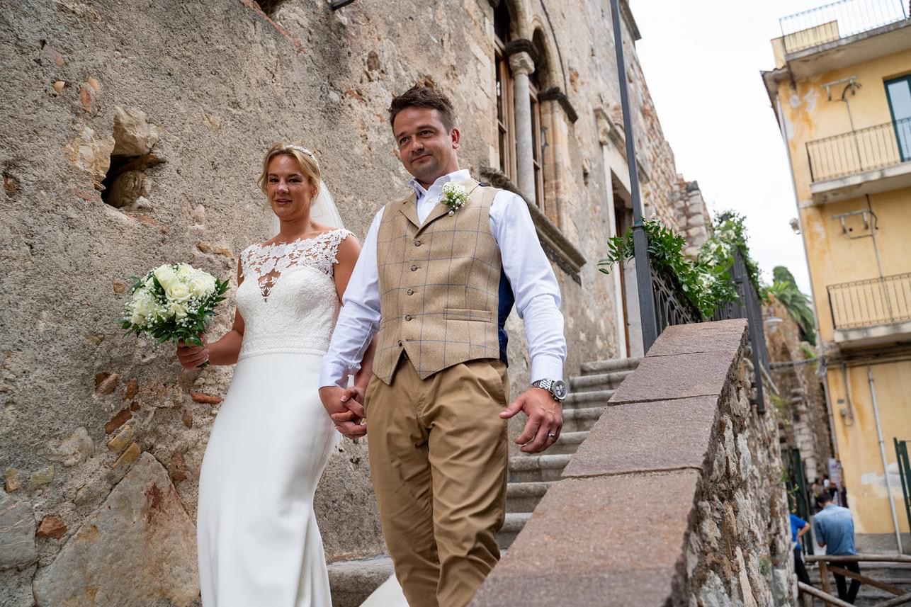 Bride and groom walking down weathered stone steps in a Sicilian hill town, the bride holding a white bouquet
