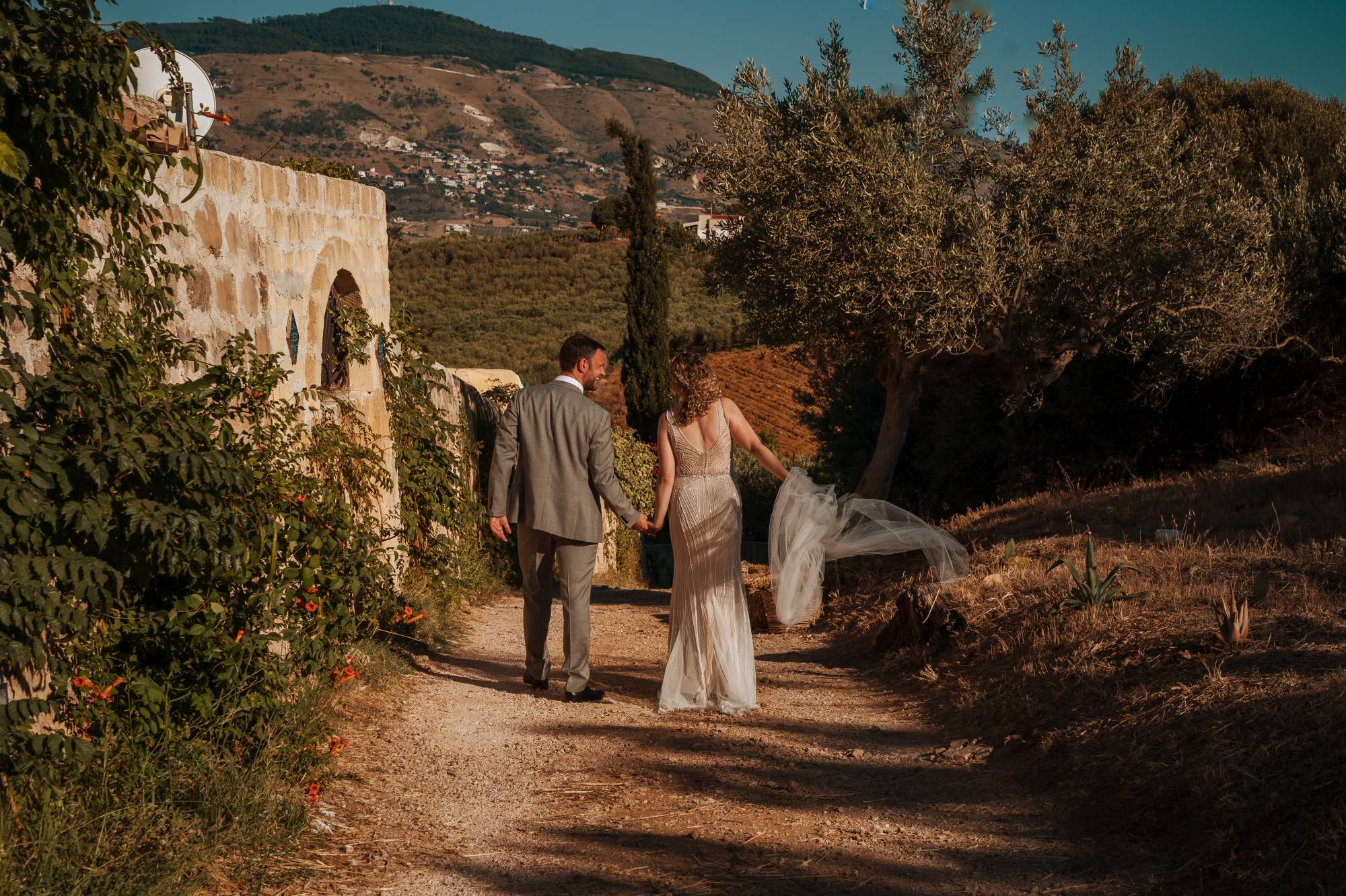 Bride and groom walking hand in hand along a country path with olive groves and rolling Sicilian hills beyond