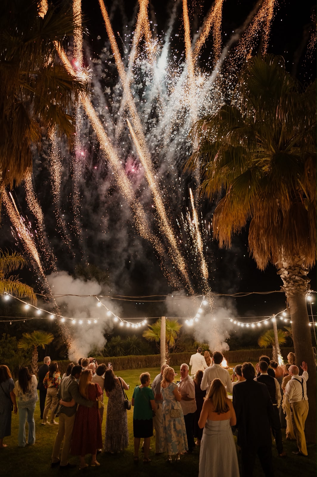 Guests watching a dazzling fireworks display over palm trees and festoon lights at a Sicilian wedding reception at night