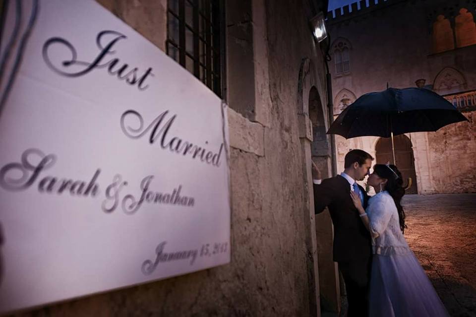 Bride and groom sharing a kiss under an umbrella beside a Just Married sign in a floodlit Sicilian piazza at night