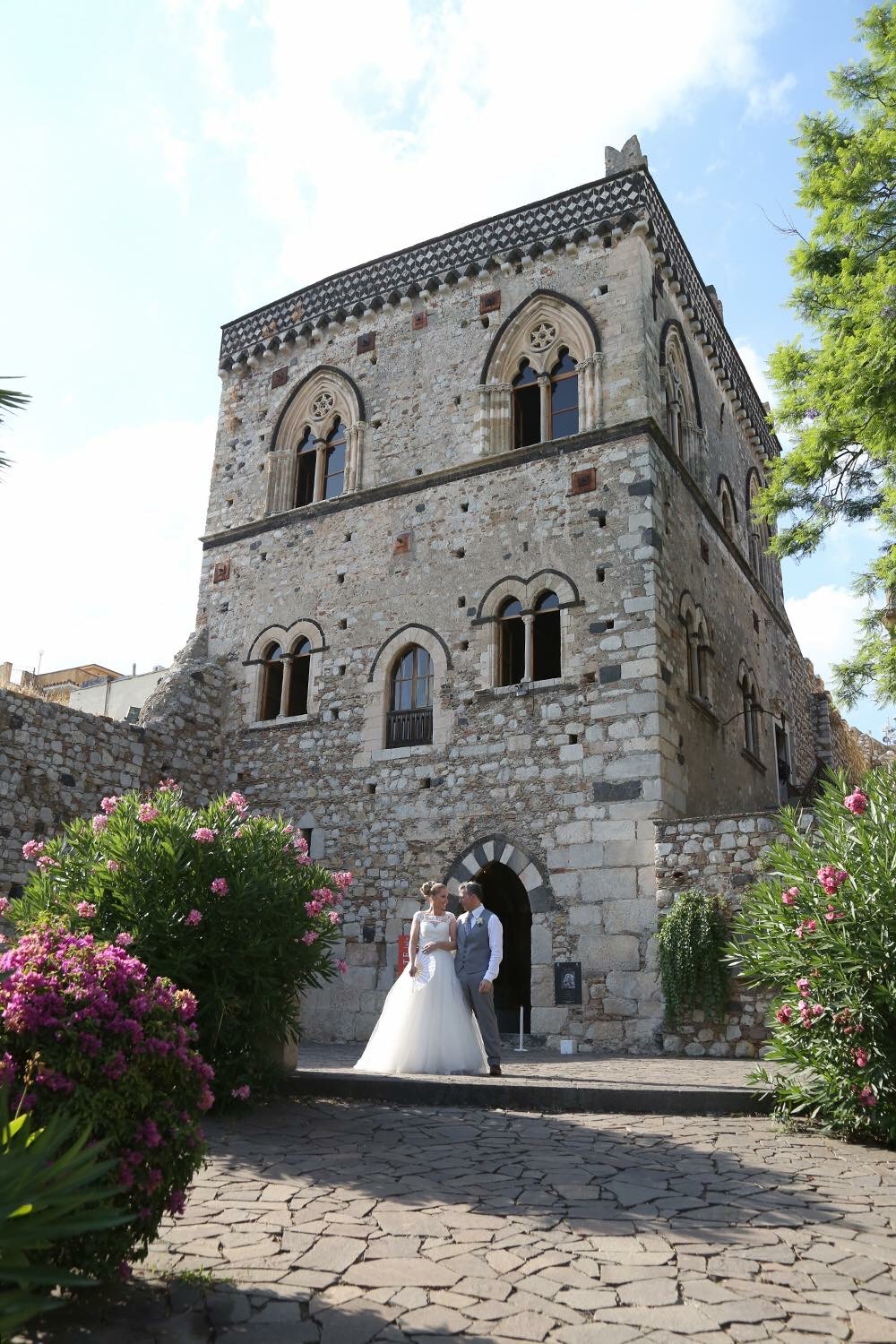 Bride and groom standing before a historic medieval stone palazzo tower framed by pink oleander blossoms in Taormina