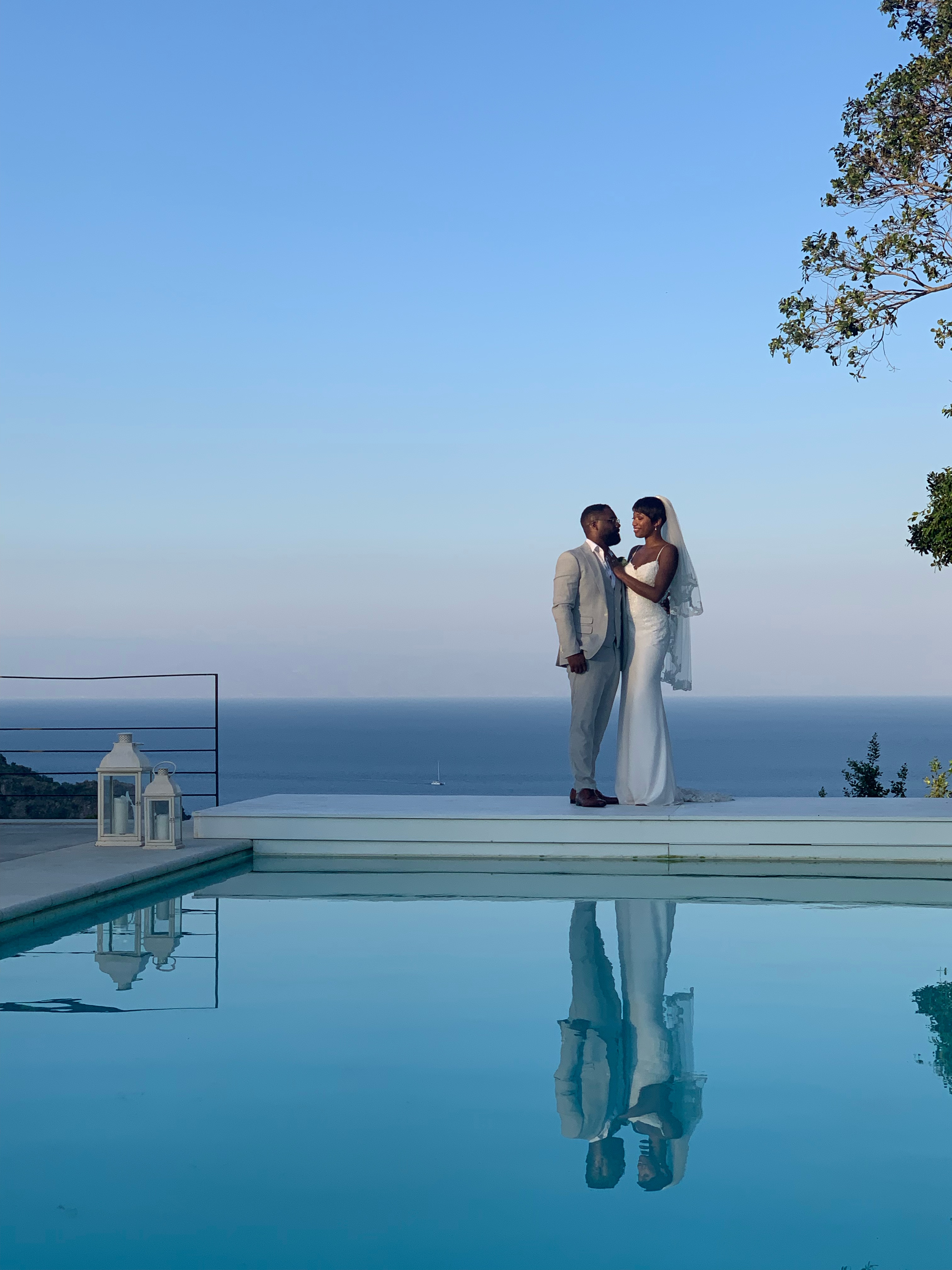 Newlyweds embracing at the edge of an infinity pool with their reflection and a sweeping Sicilian sea view at dusk