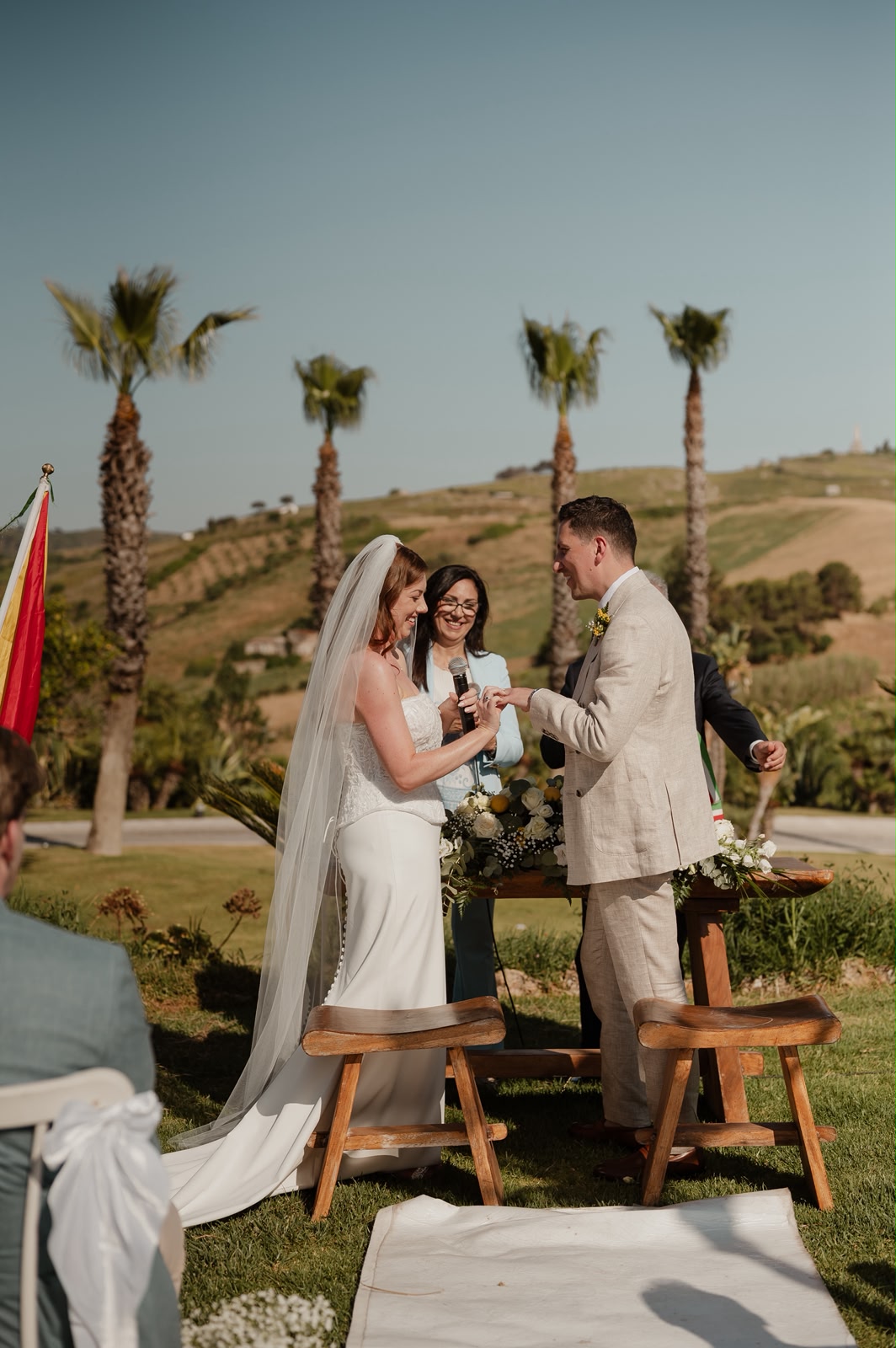Bride and groom exchanging rings with their celebrant during an outdoor ceremony framed by palm trees and rolling Sicilian hills
