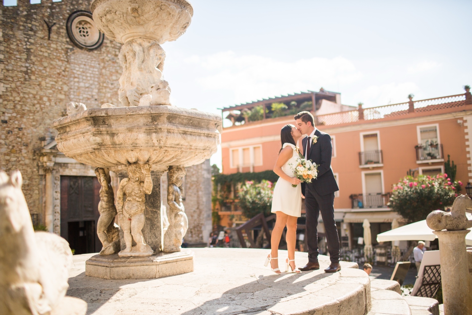 Bride and groom kissing beside a baroque stone fountain in a sunlit Taormina square