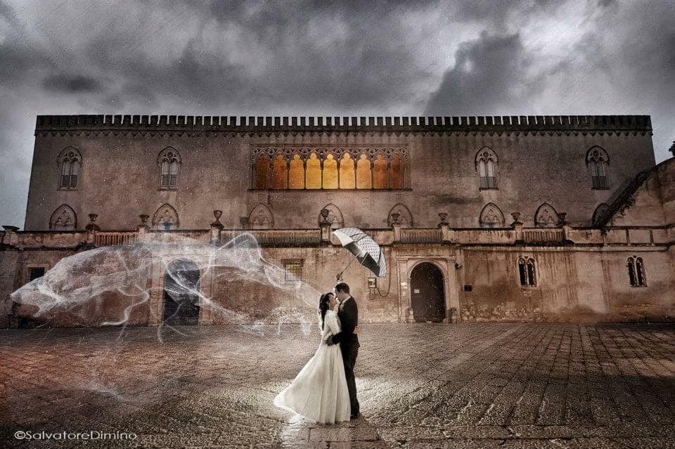 Dramatic evening portrait of a couple embracing beneath a parasol in a historic Sicilian palazzo courtyard with a flowing veil