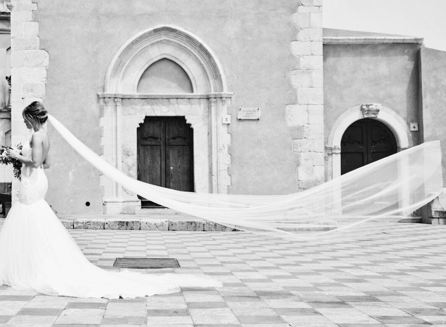 Black and white photograph of a bride with a dramatic cathedral-length veil sweeping across a historic Taormina piazza
