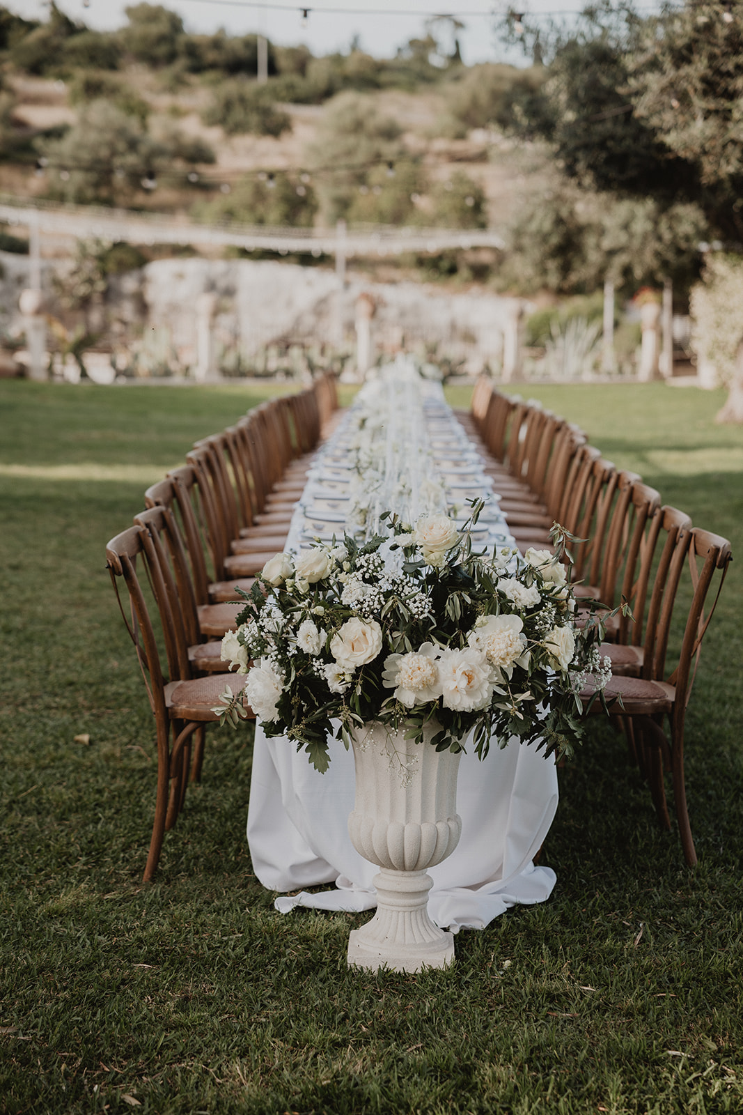 The long alfresco imperial table on the lawn, crowned with white flowers in a stone urn