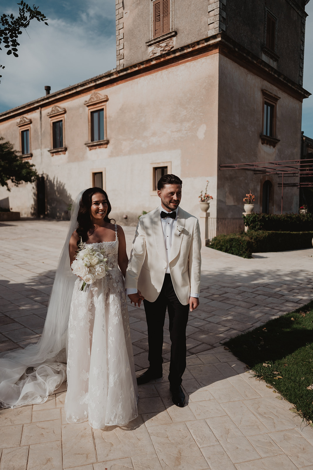 Lidia and Rian hand in hand in the sunlit courtyard of Borgo del Carato, Sicily