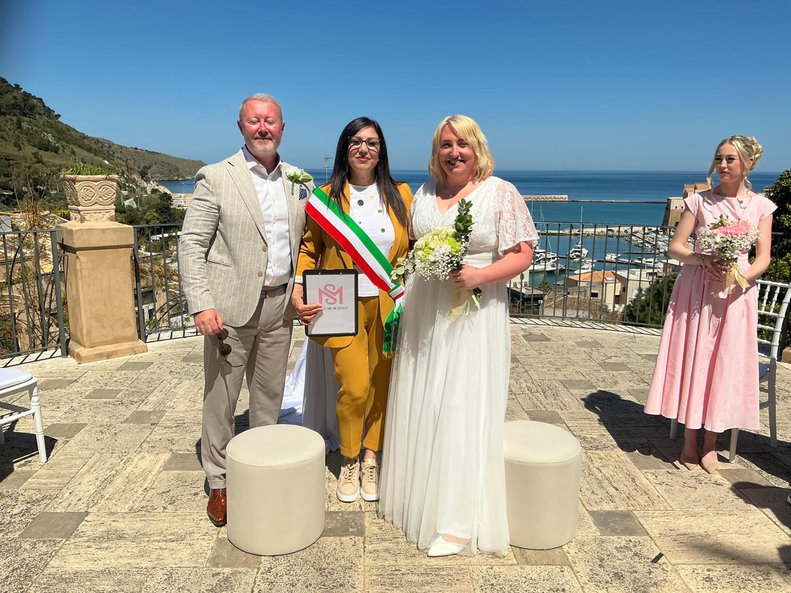 Sabina, wedding celebrant, wearing the Italian tricolour sash and holding the Marry Me in Sicily folder, beside a married couple at a Sicilian harbour
