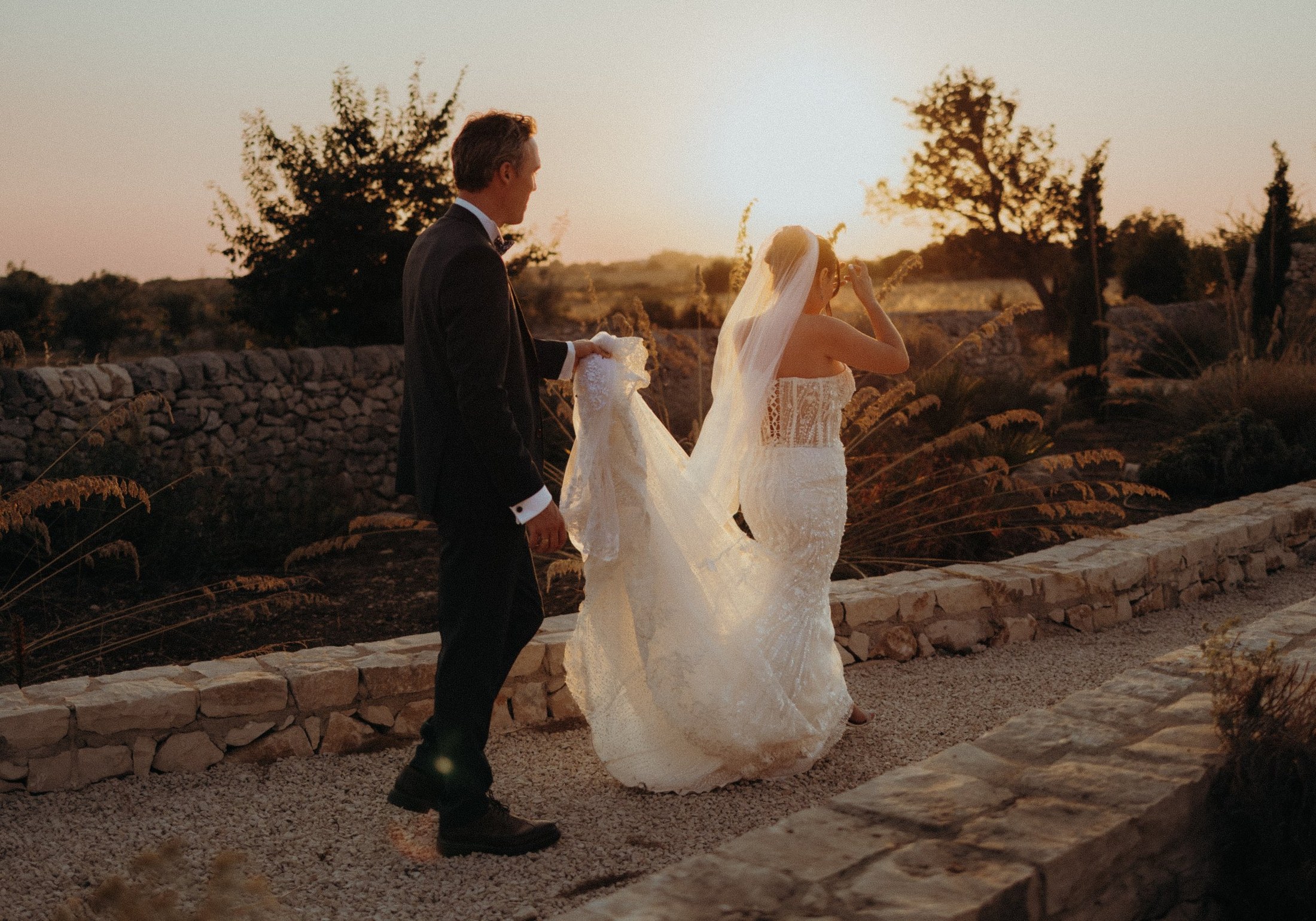 Sicilian wedding couple walking a gravel garden path in golden evening light past low dry-stone walls and open countryside