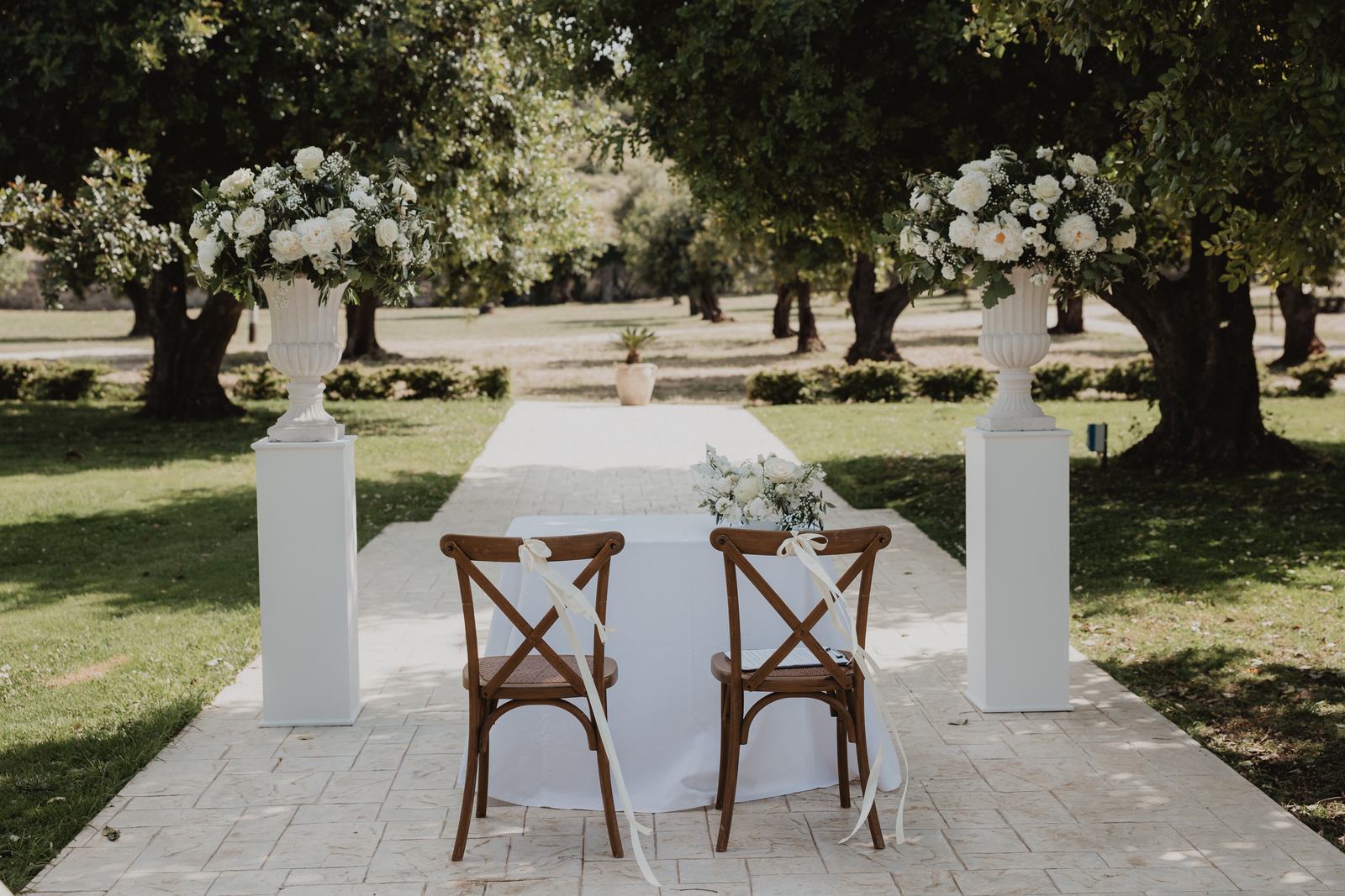 A wedding ceremony table set beneath the trees in the gardens of Borgo del Carato near Siracusa, with white flowers on stone pedestals