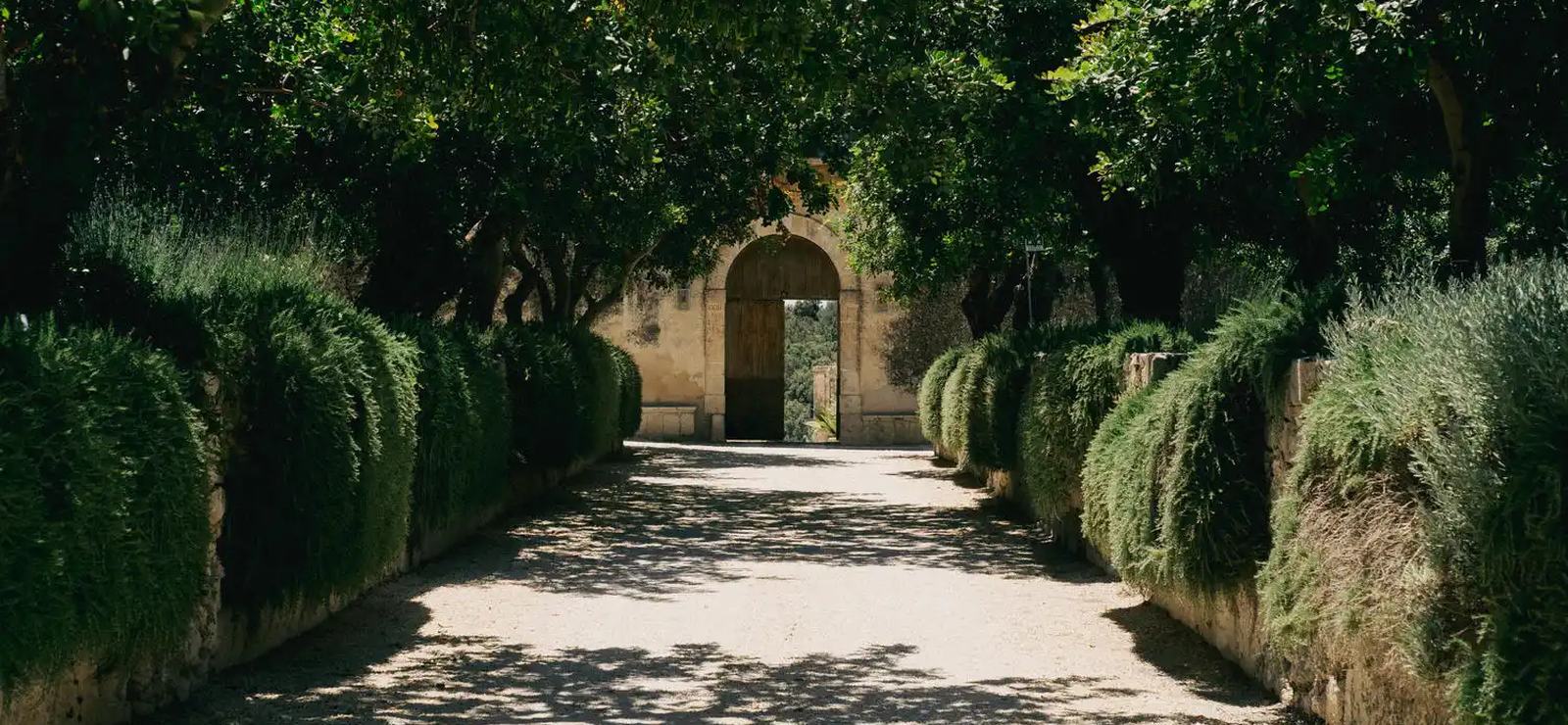 A tree-lined garden path leading to an arched gateway at Dimora delle Balze, a boutique estate in Sicily
