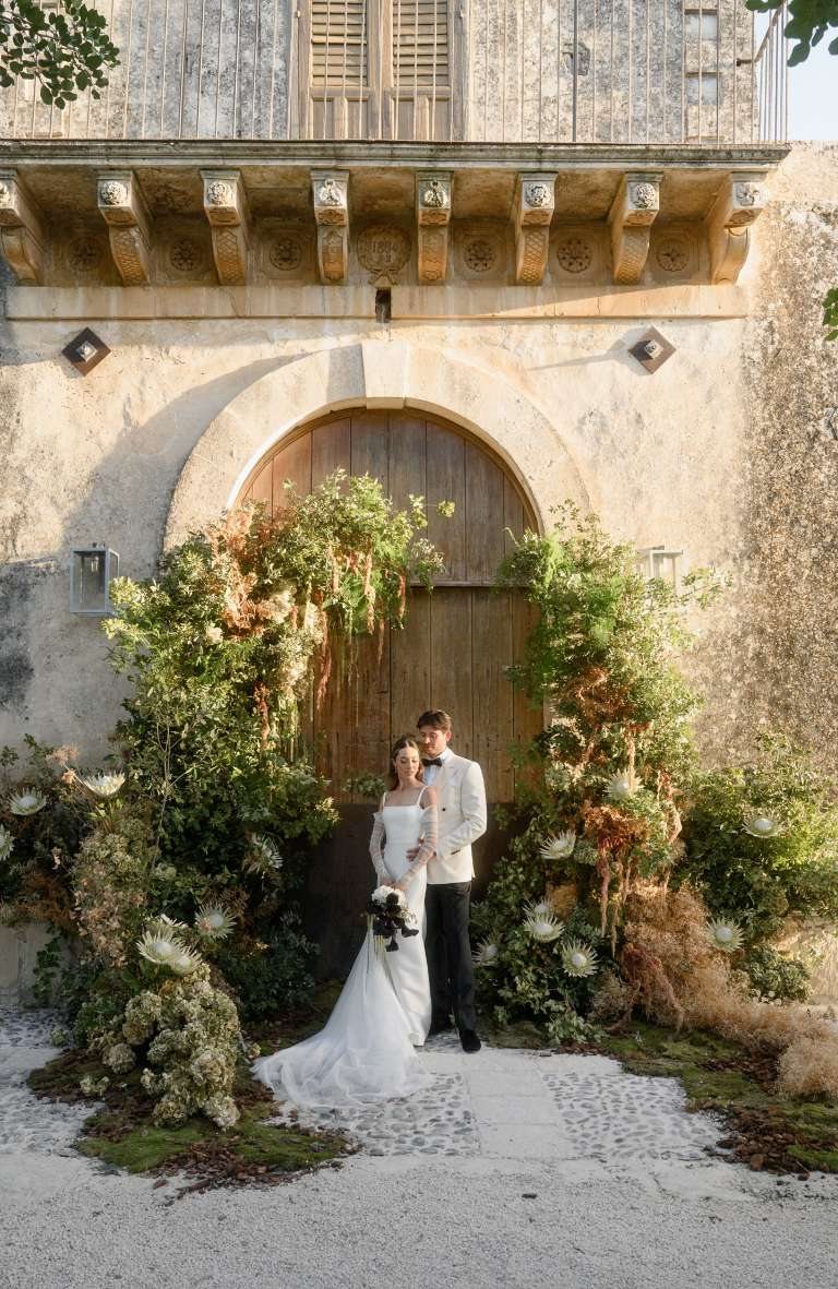 Bride and groom in front of the old arched manor door framed by a flower installation at Dimora delle Balze