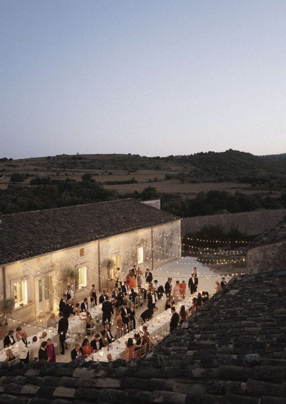 Wedding guests at a long dinner table beside the masseria at sunset, with the Val di Noto hills beyond
