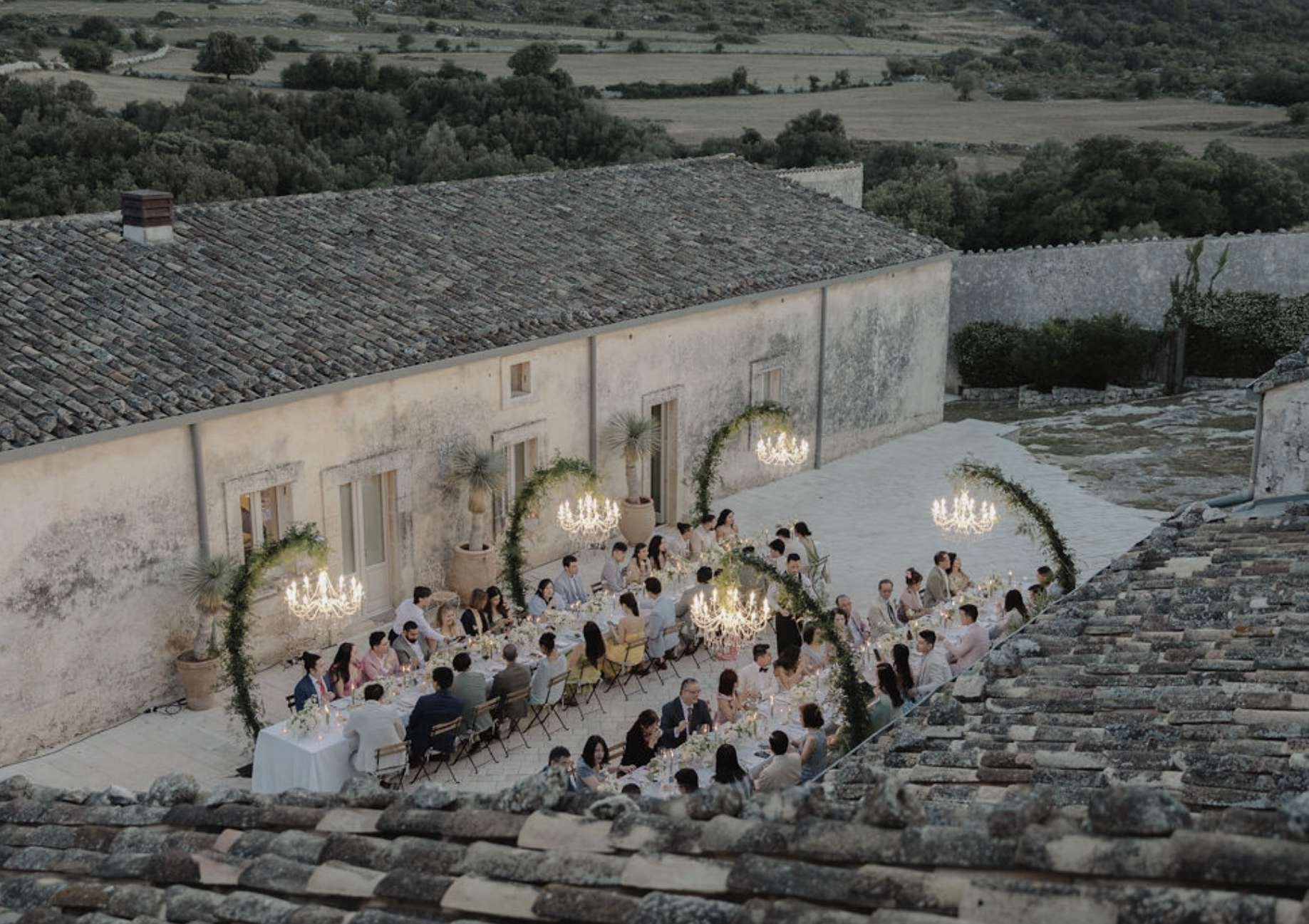 Wedding dinner with floral hoops and chandeliers beside the old stone masseria at dusk, seen from the rooftops