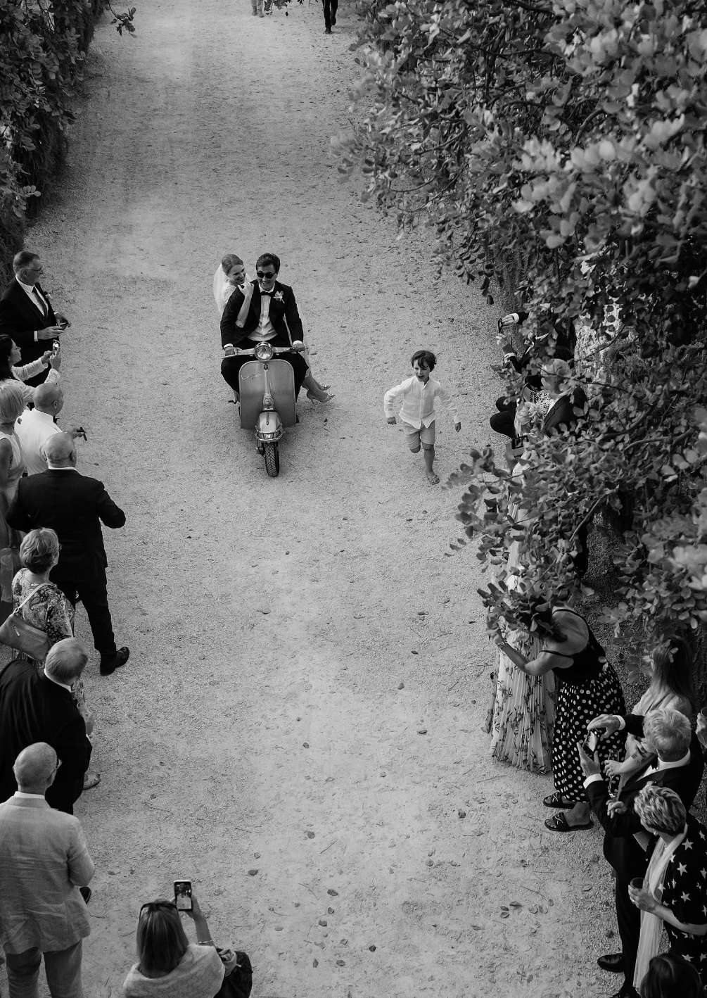Black-and-white photo of newlyweds riding a scooter down the Viale dei Carrubi as guests cheer at Dimora delle Balze