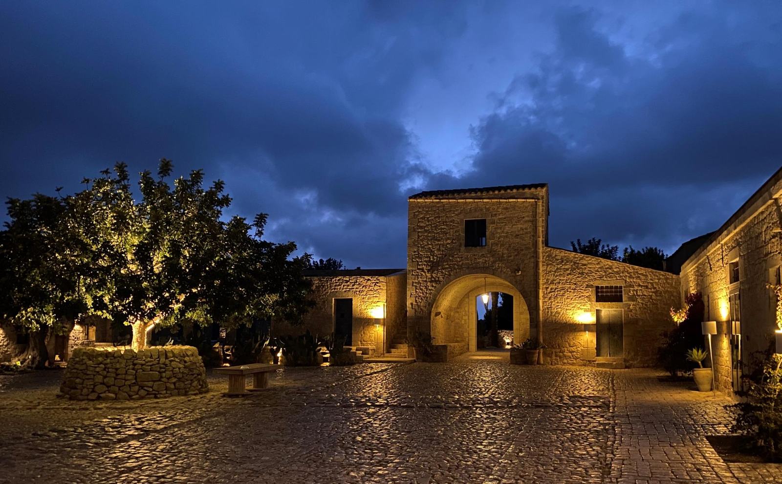 Il cortile in pietra de La Chiusa Country House illuminato al tramonto, con l’ingresso ad arco del casale e gli ulivi